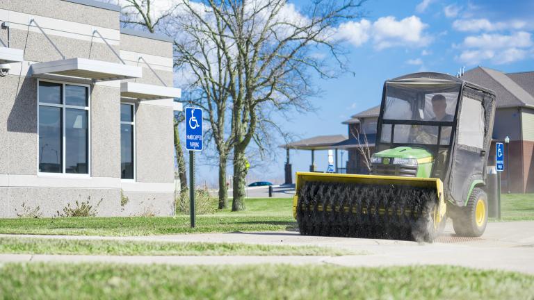 a riding mower with a sweeper attachment cleaning the sidewalk outside of an office building